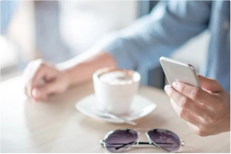 A person is holding a smartphone with their right hand while sitting at a table with a cup of coffee and sunglasses.
